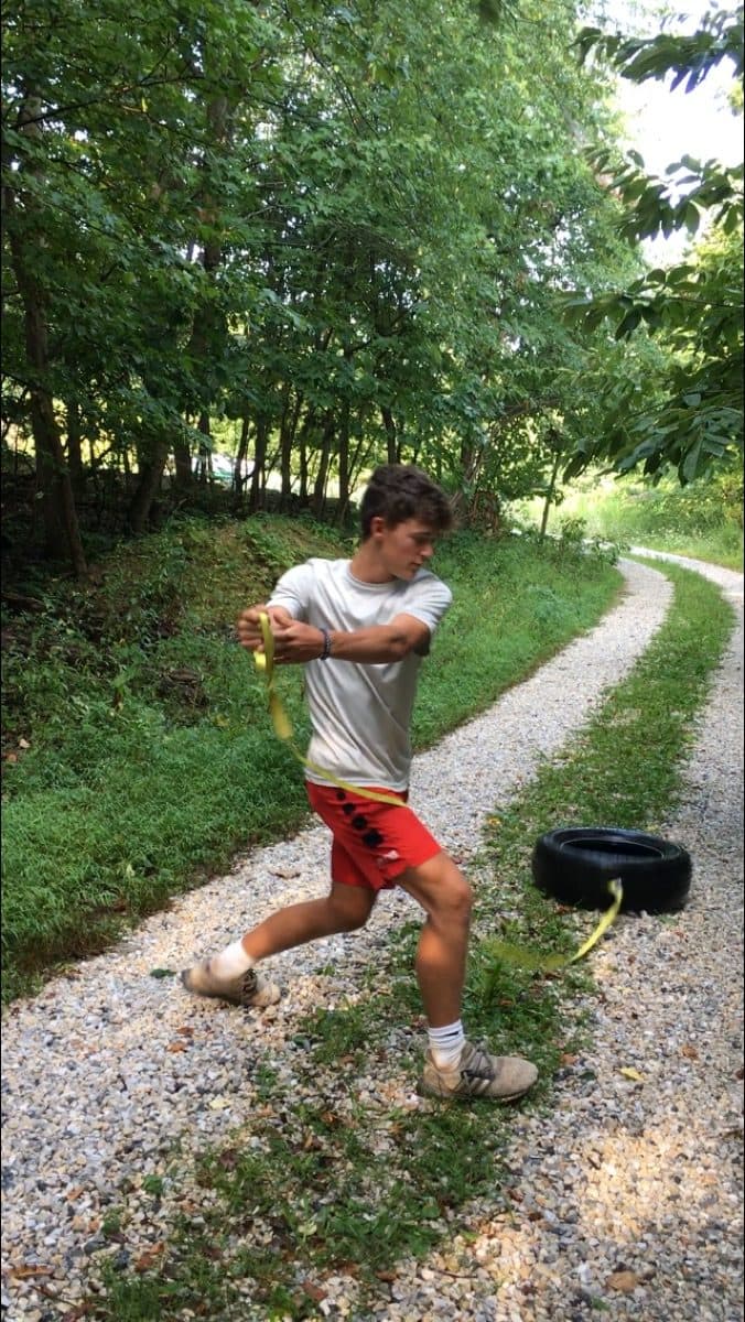A young athlete performing a lunge with rotation while towing a weighted tire along a gravel lane.