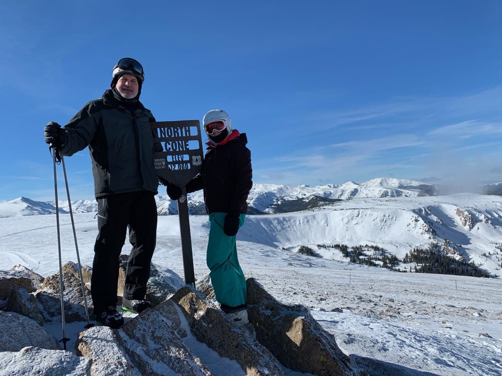 Two skiers standing beside the North Cone summit sign at 12,060 feet, snow-covered Colorado peaks behind them.